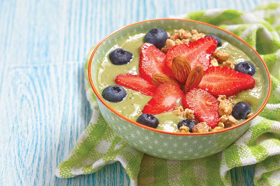 Green coloured smoothie in a patterned bowl covered in fruit and nuts.