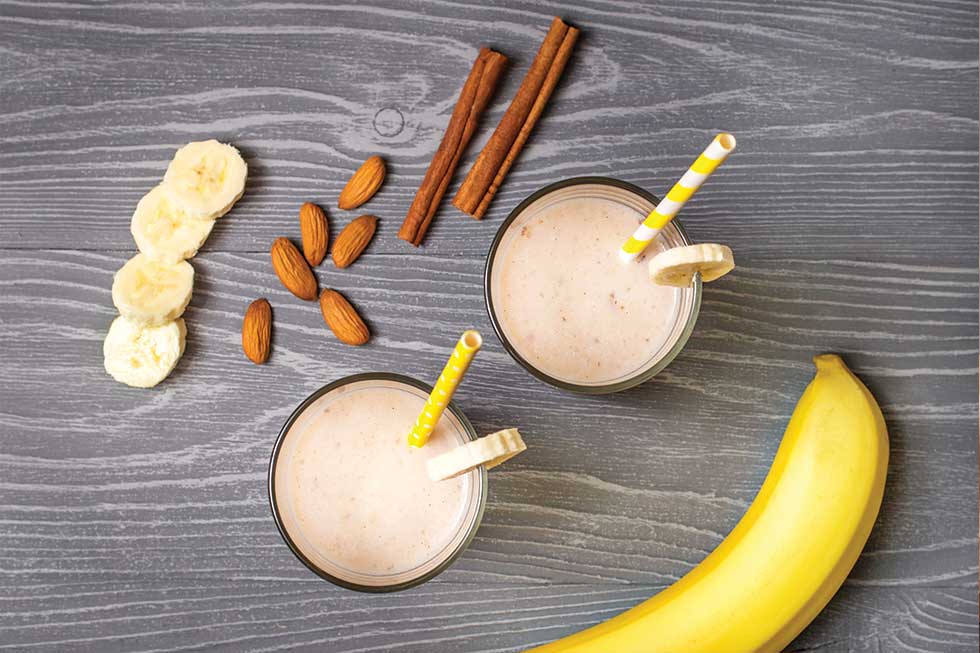 Two glasses of light brown protein smoothie with yellow paper straws and bananas, cinnamon and almonds surrounding it.